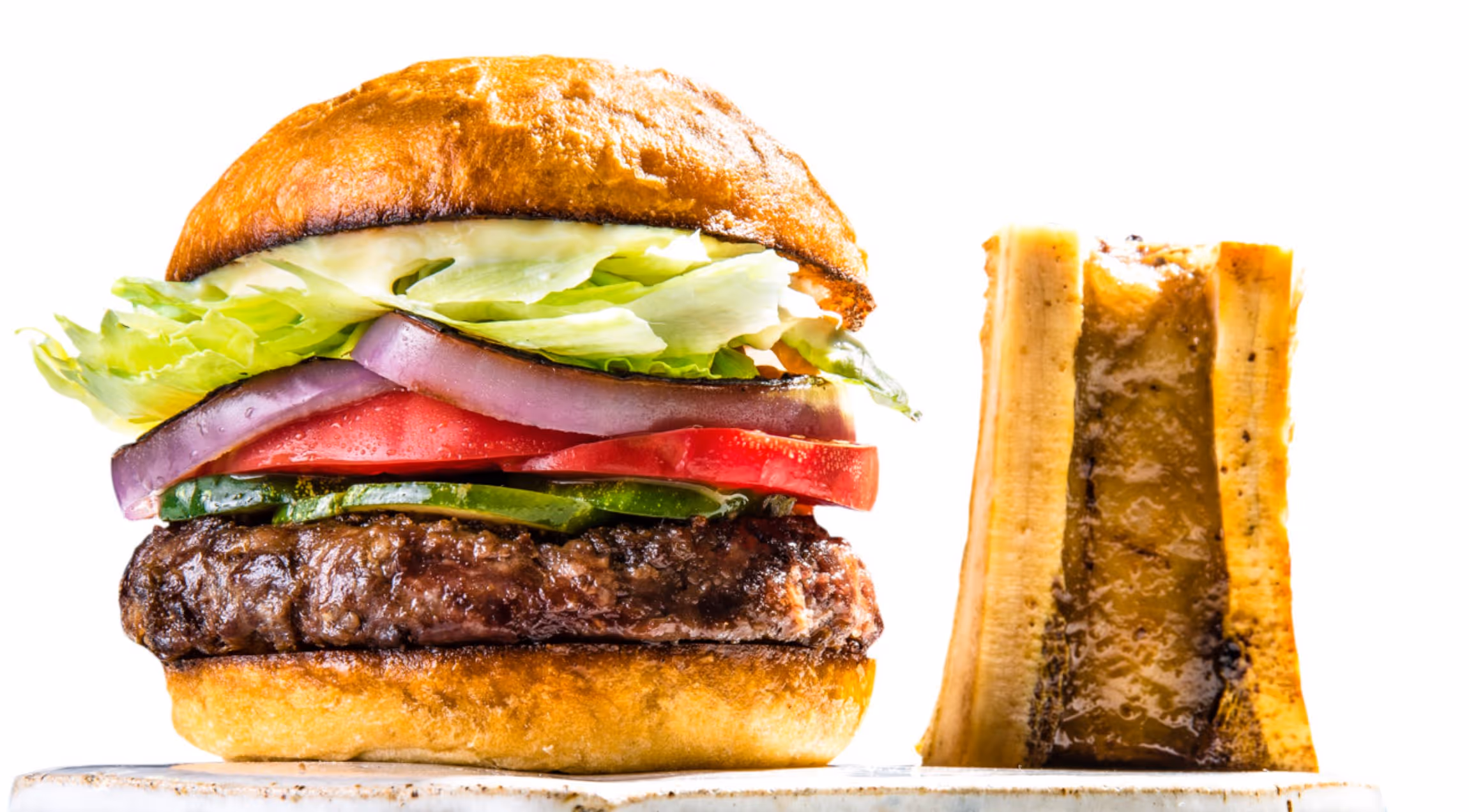 Grilled hamburger with lettuce, red onion, tomato, and cucumber on a toasted bun beside a grilled banana half.