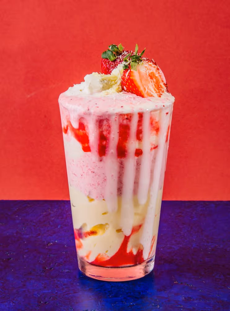 Glass of creamy strawberry milkshake topped with whipped cream and fresh strawberries against a red and blue background.