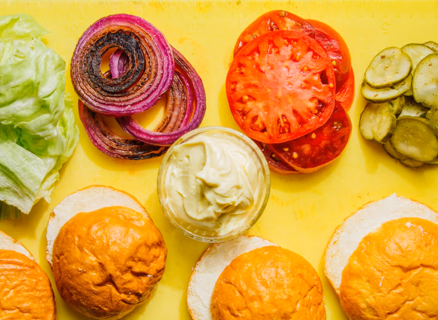 Burger buns, iceberg lettuce, grilled red onion rings, tomato slices, pickles, and a glass bowl of creamy sauce arranged on a yellow surface.