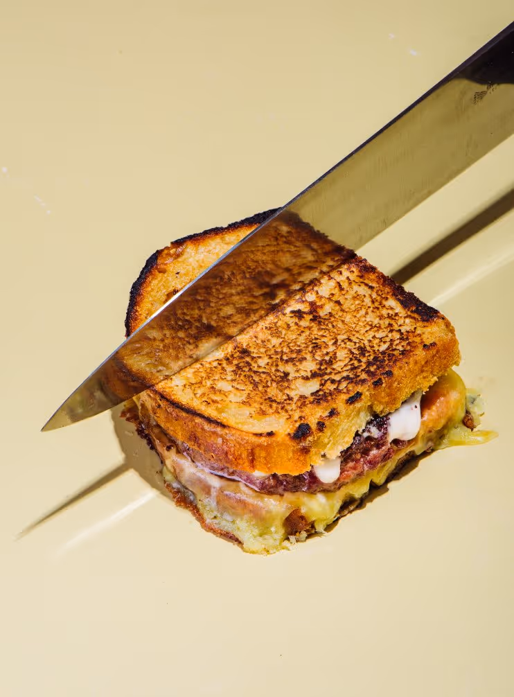 Grilled cheese sandwich with melted cheese and meat being sliced by a large knife on a beige plate.