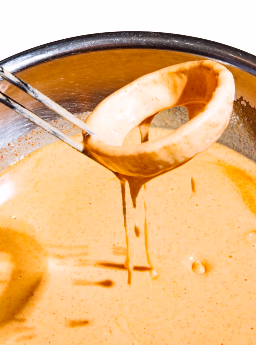 Metal ladle lifting a ring-shaped food item coated in creamy orange batter over a bowl of the same batter.