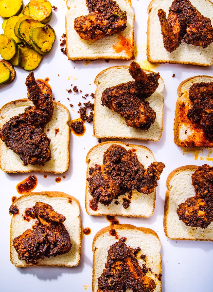 Spicy fried chicken wings placed on slices of white bread with a side of grilled zucchini slices on a white surface.