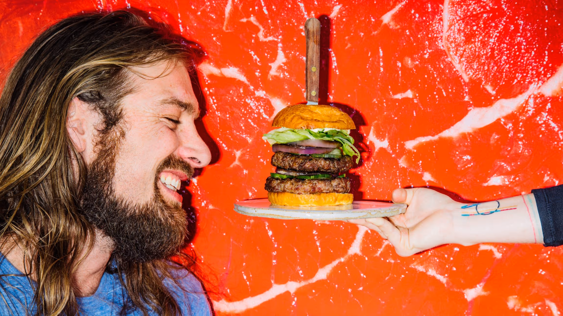 Bearded man with long hair smiling at a double cheeseburger with lettuce and onion, held on a plate by a tattooed hand, against a red marbled background.