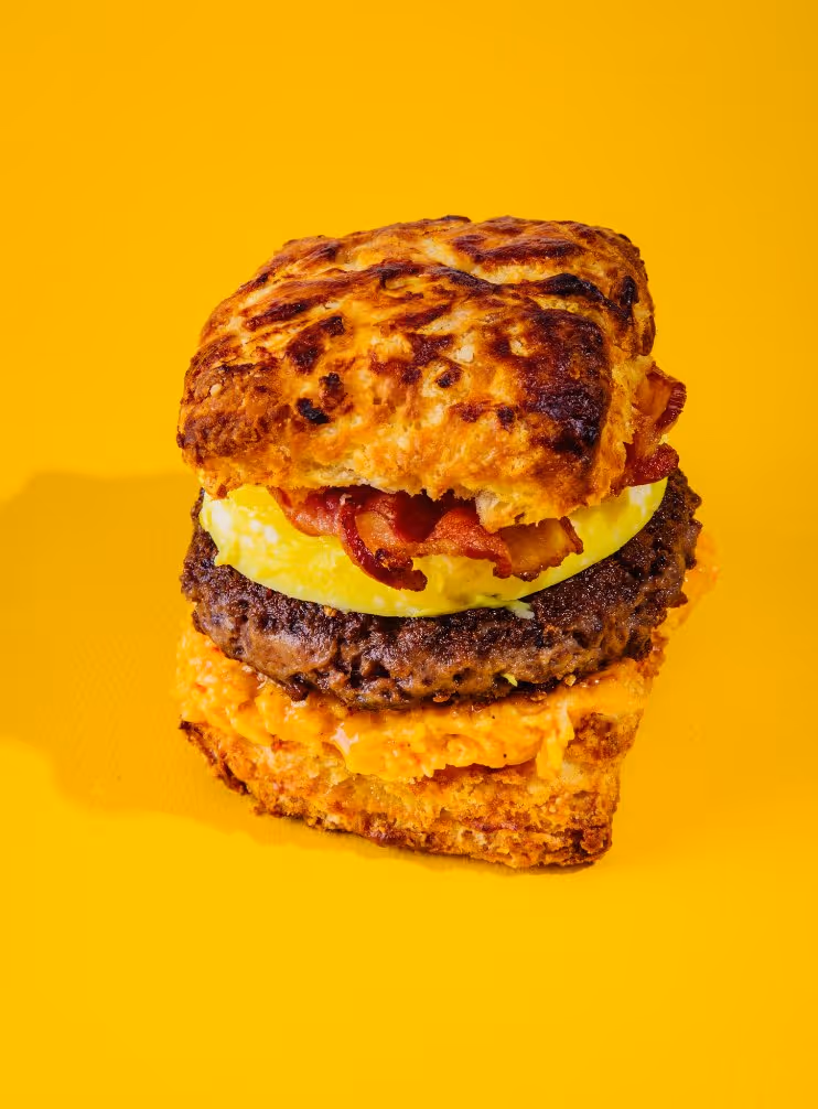 Square biscuit sandwich with crispy bacon, egg, beef patty, and cheese spread on a yellow background.