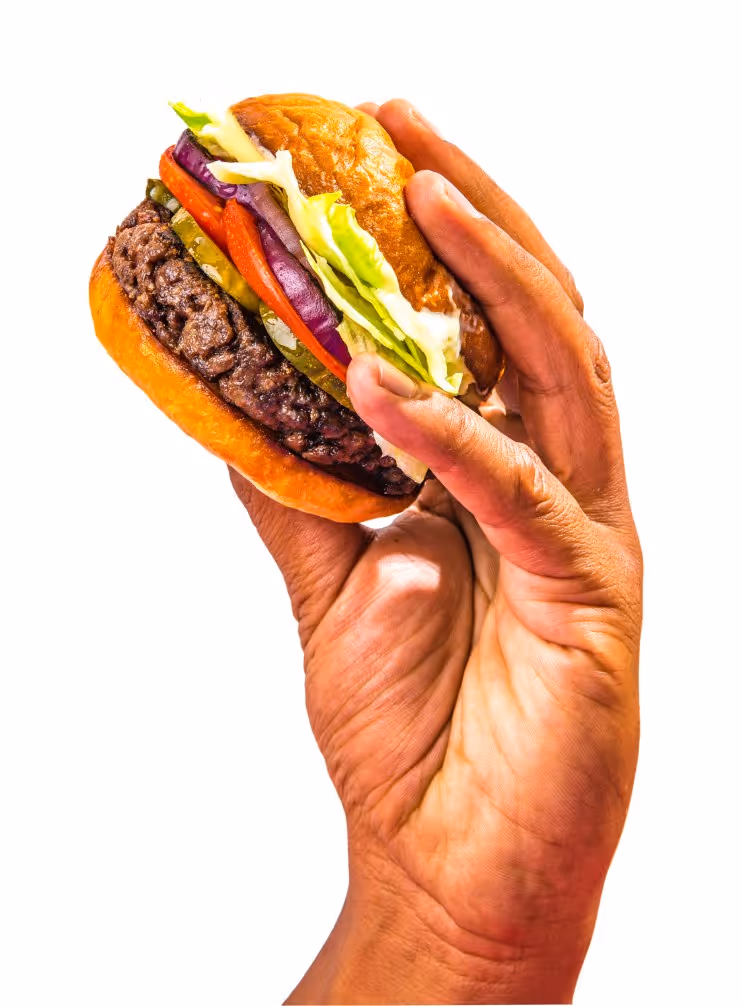 Hand holding a hamburger with lettuce, tomato, pickles, red onion, and beef patty in a bun.