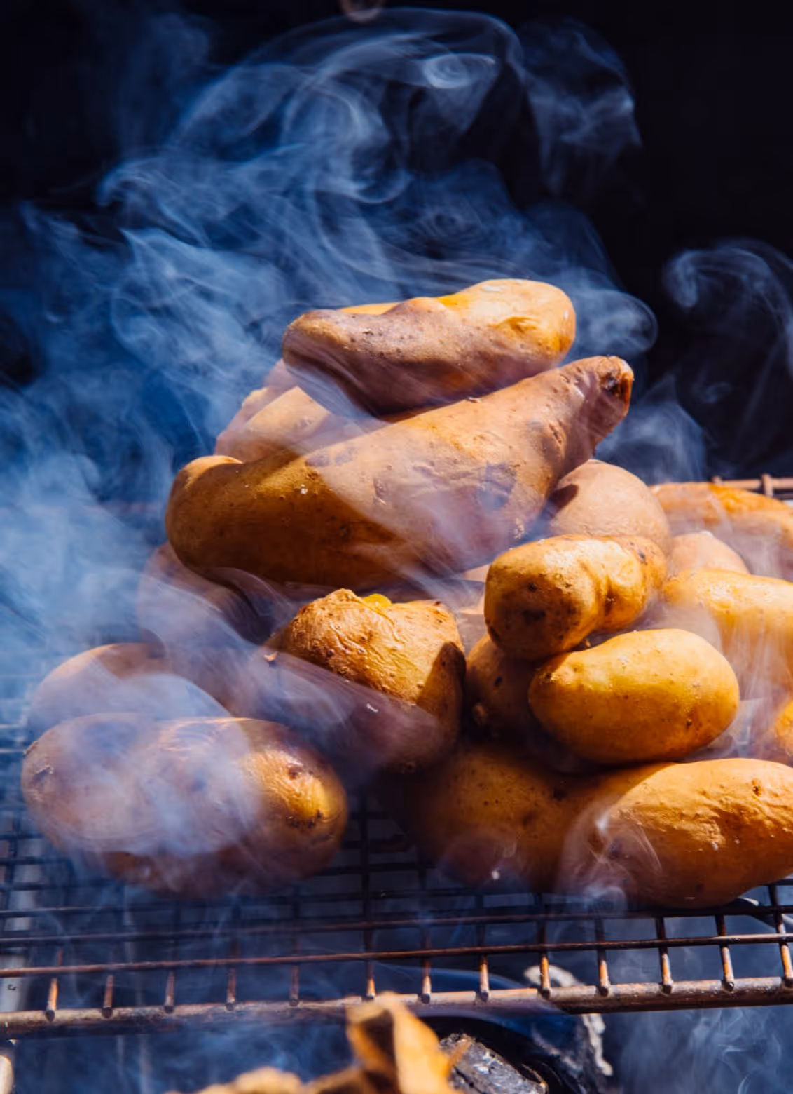 Sweet potatoes roasting on a grill with smoke rising around them.