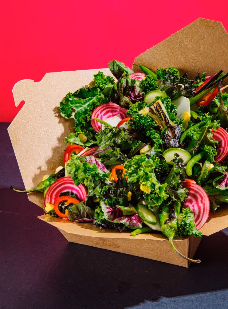 Fresh mixed salad with kale, striped beet slices, cucumber, red bell pepper, and grilled vegetables in a cardboard takeout container.