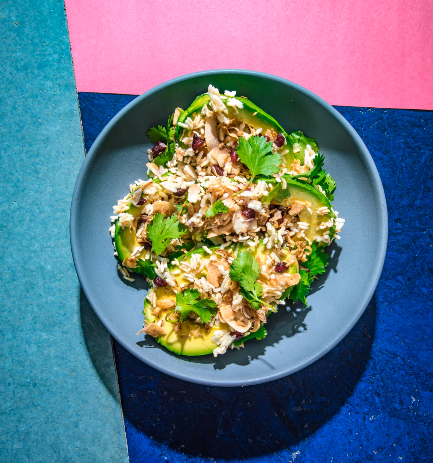 Gray plate with avocado slices topped with puffed rice, herbs, and dried berries on a colorful blue and pink background.