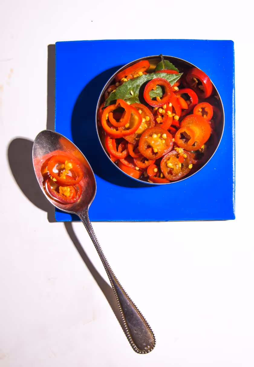 Sliced red chili peppers with seeds in a round metal bowl on a blue square plate and a spoon holding some chili slices.