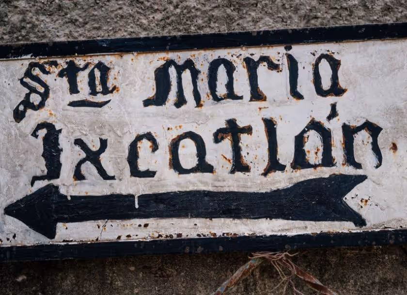 Rustic white sign with black Gothic-style text 'sta maria Ixcatlán' and a black arrow pointing left.