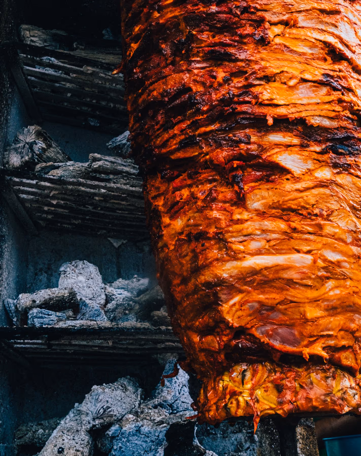 Close-up of marinated meat roasting on a vertical spit beside burning charcoal.