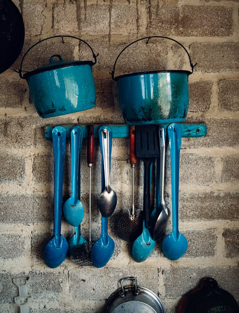 Two blue metal pots hanging on a rough brick wall above a row of various blue and metal kitchen utensils.