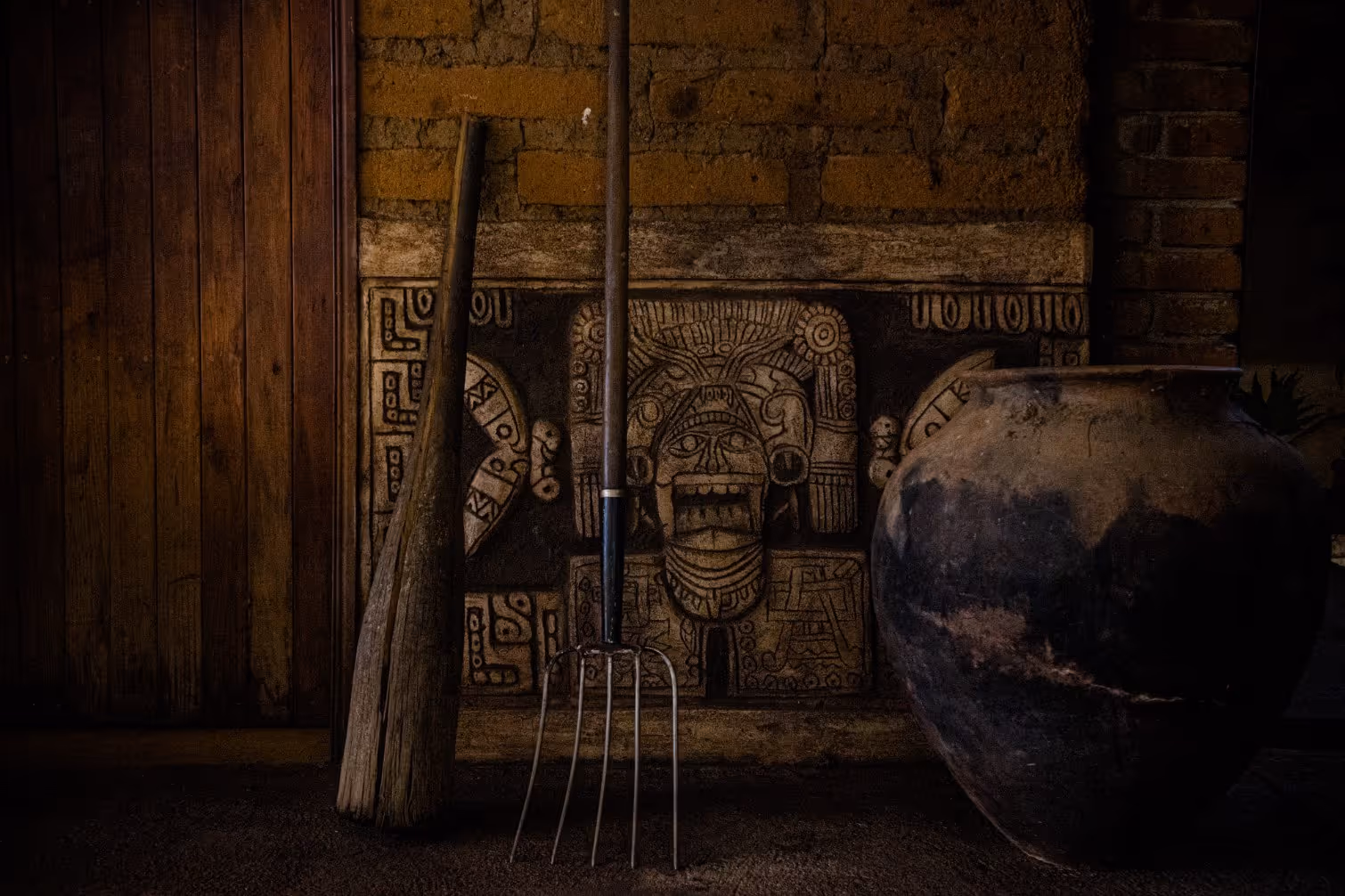 Rustic wooden broom and metal garden fork standing in front of a stone carving with an ancient face design and a large clay pot beside them.