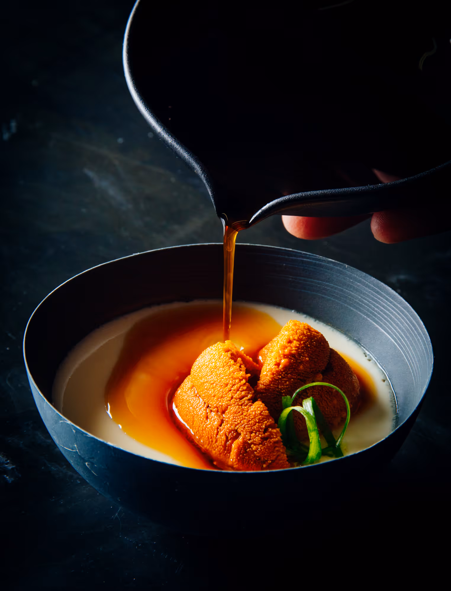 Orange sea urchin in creamy soup with green garnish being drizzled with brown broth from a black pouring vessel.