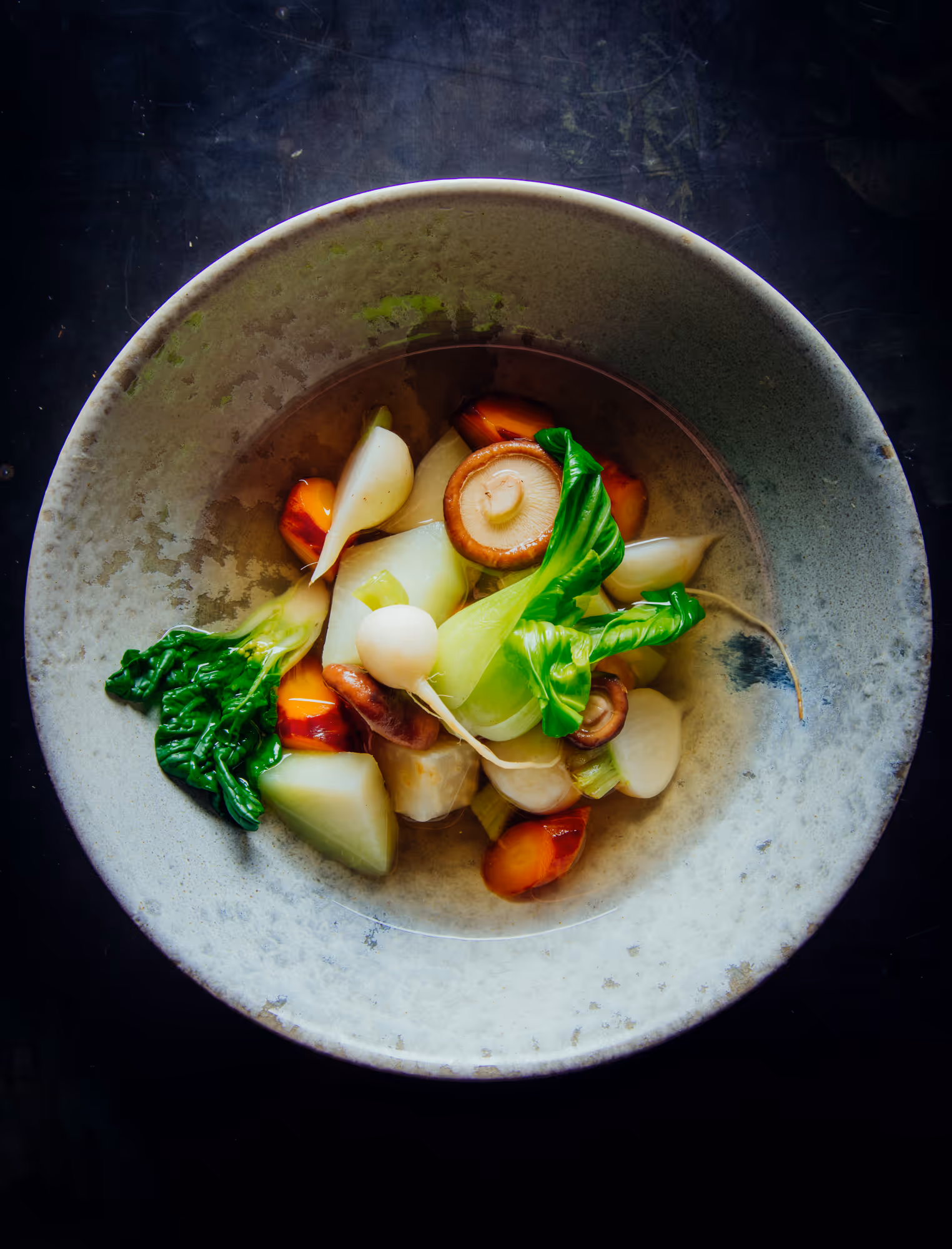 Bowl of clear vegetable broth with bok choy, mushrooms, turnips, carrots, and green leaves.