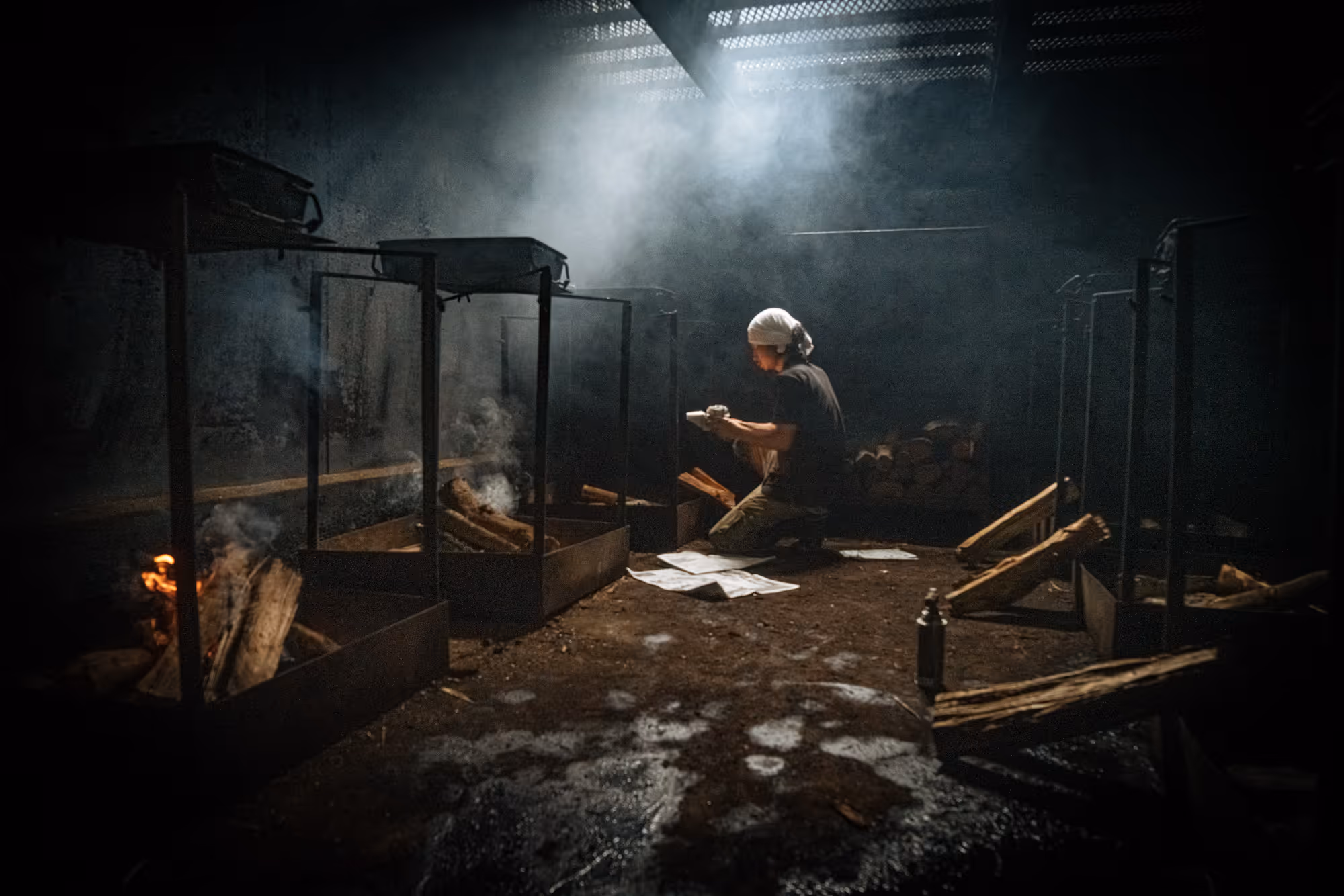 Person kneeling indoors tending to wood fires in metal fire pits with smoke rising and sunlight filtering through a roof vent.