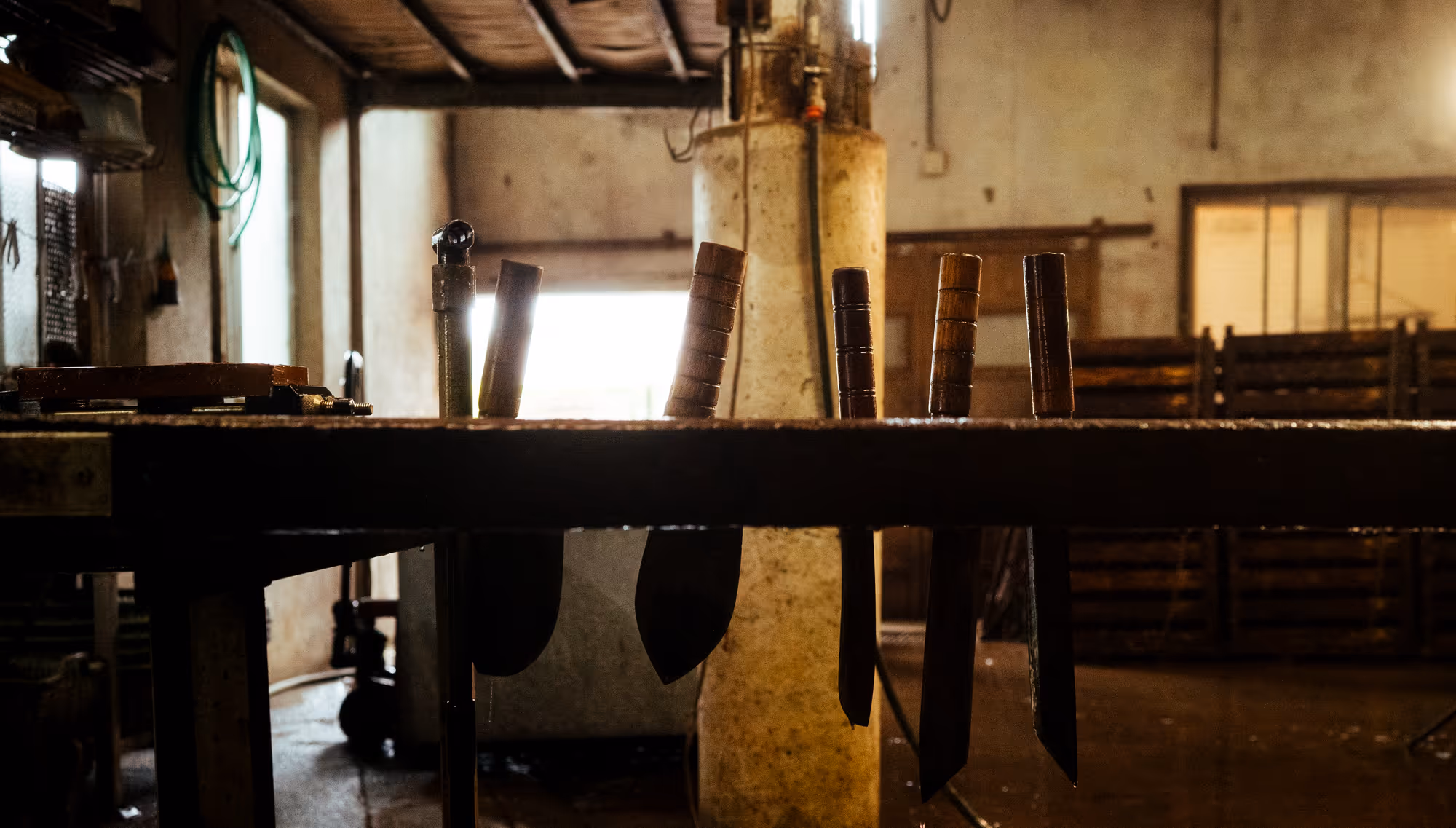 Row of machetes with wooden handles hanging from a rack in a workshop with dim lighting.