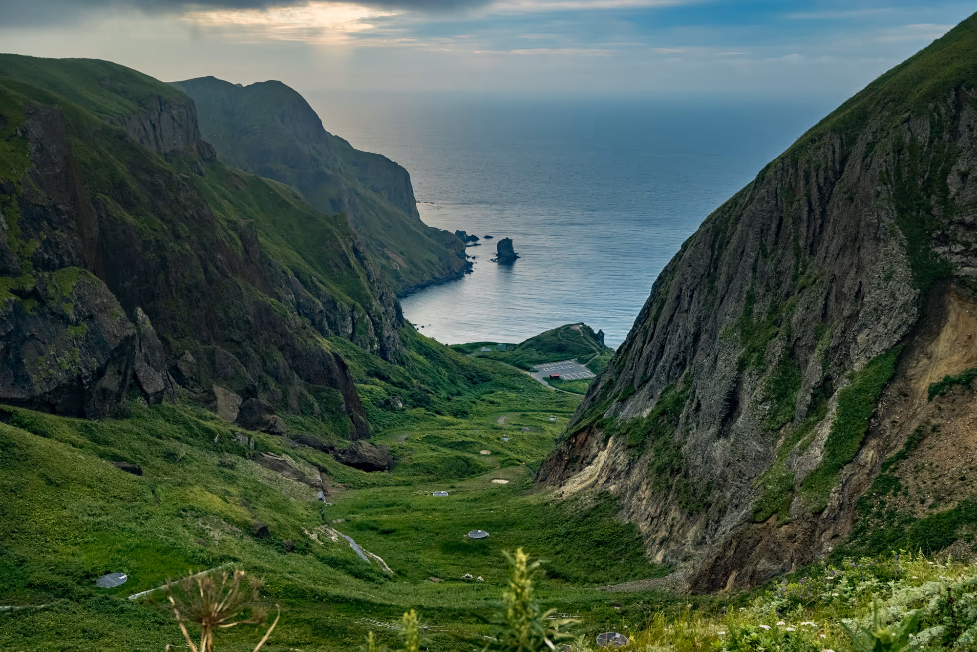 Green valley between steep cliffs leading to a rocky coastline with a calm sea under a partly cloudy sky.