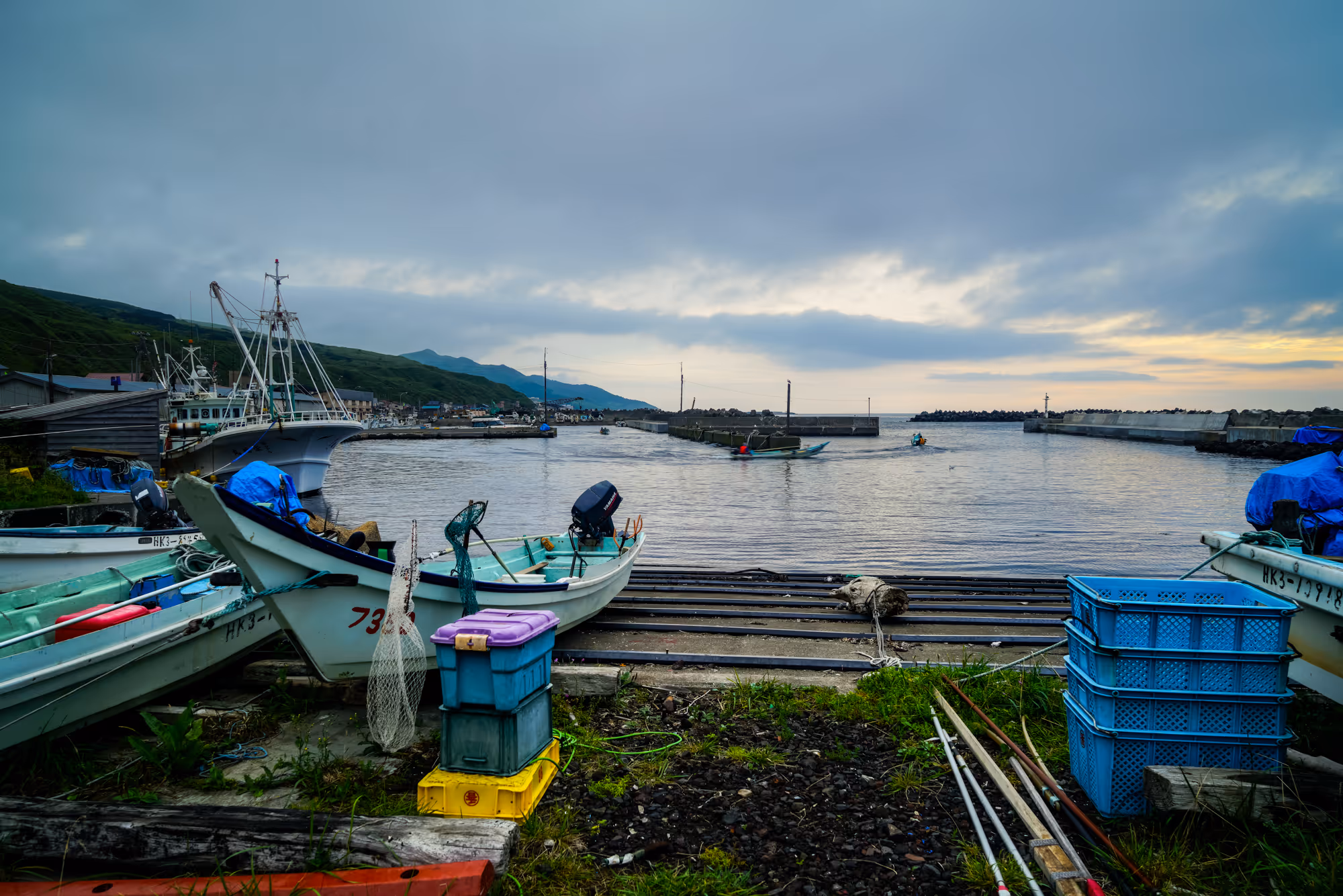 Small fishing boats docked at a harbor with stacks of crates and fishing nets, under a cloudy sky at dusk.