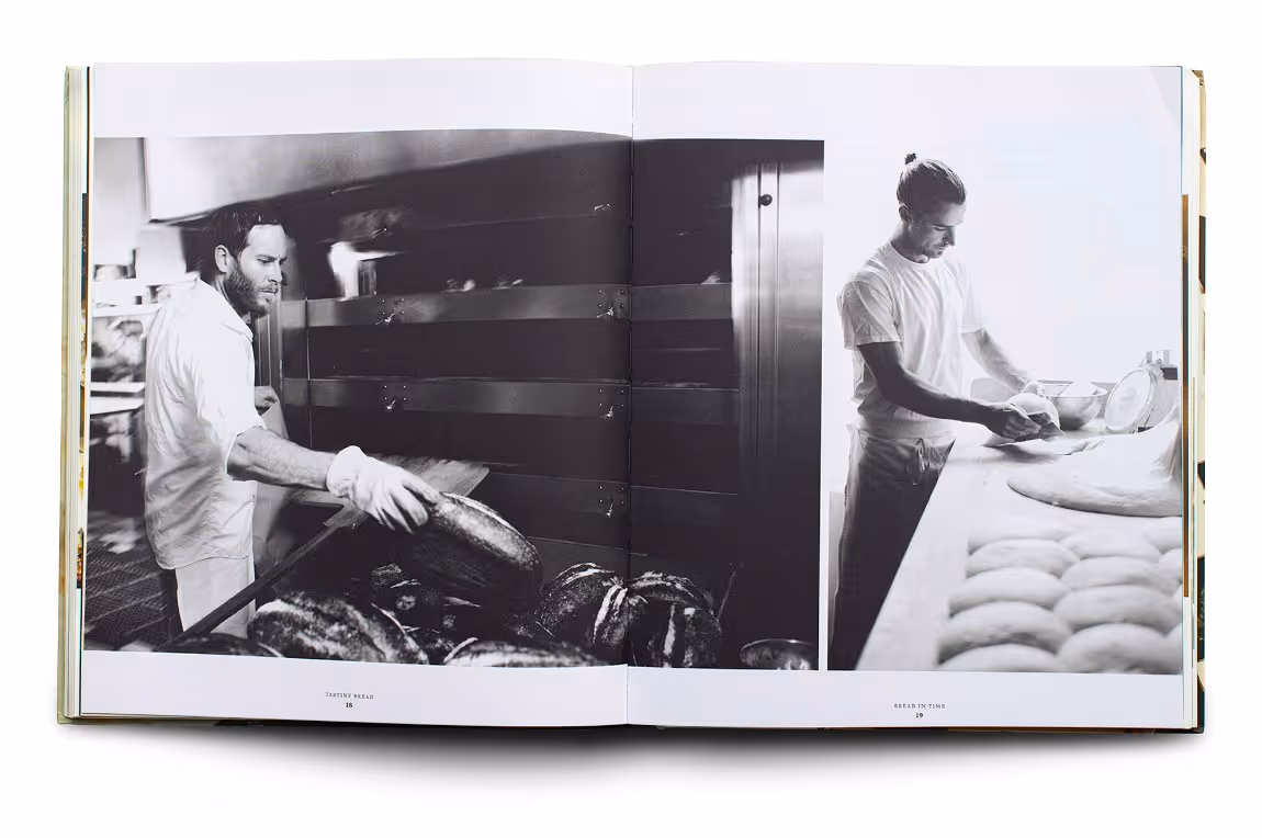 Black and white photo of a baker removing bread from an oven and another baker shaping dough at a worktable.