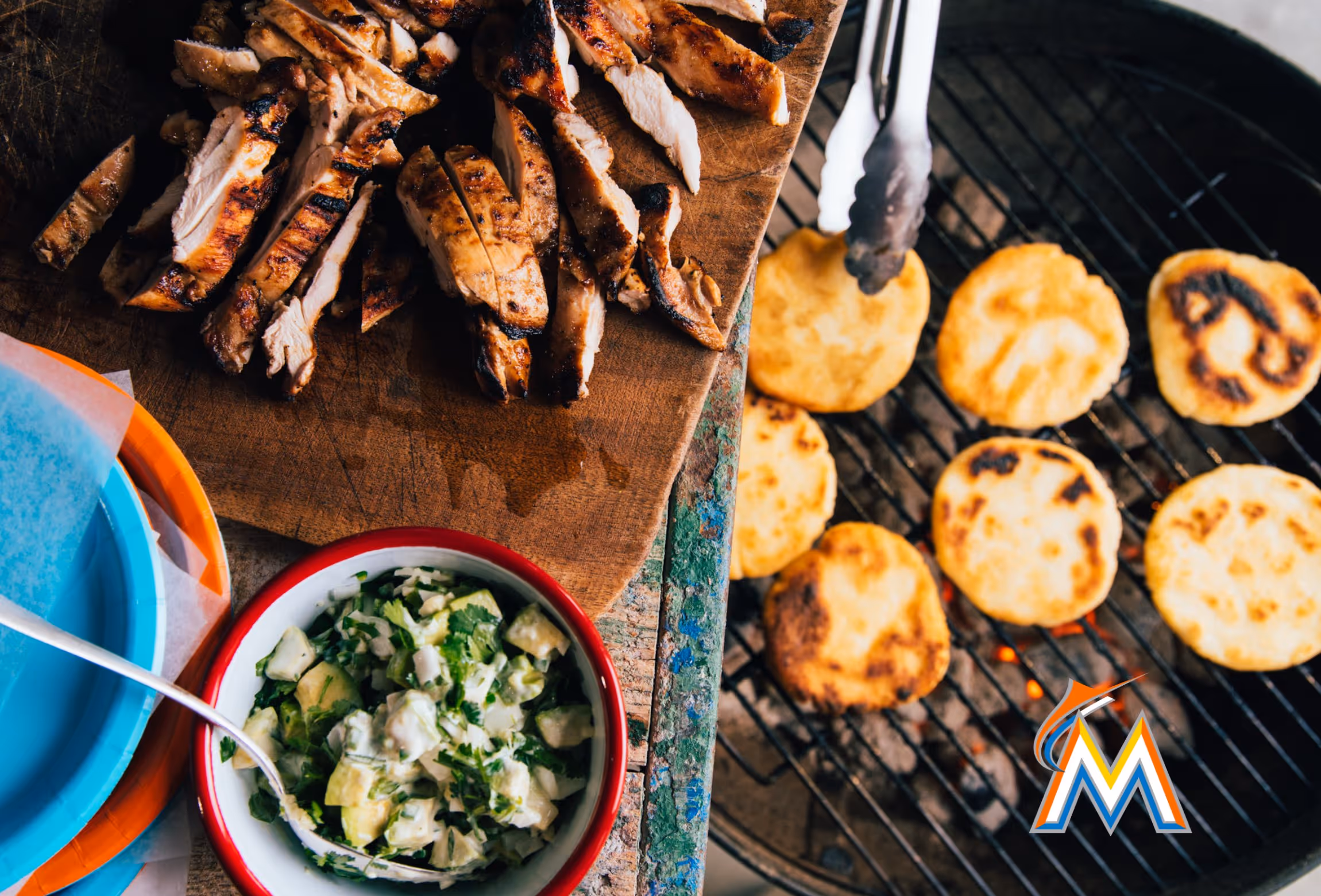 Grilled chicken slices on a wooden board with a bowl of avocado salad and arepas cooking on a grill with tongs.