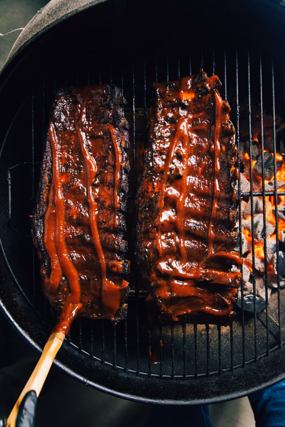 Two racks of ribs coated with barbecue sauce on a charcoal grill, with sauce being brushed on one rack.