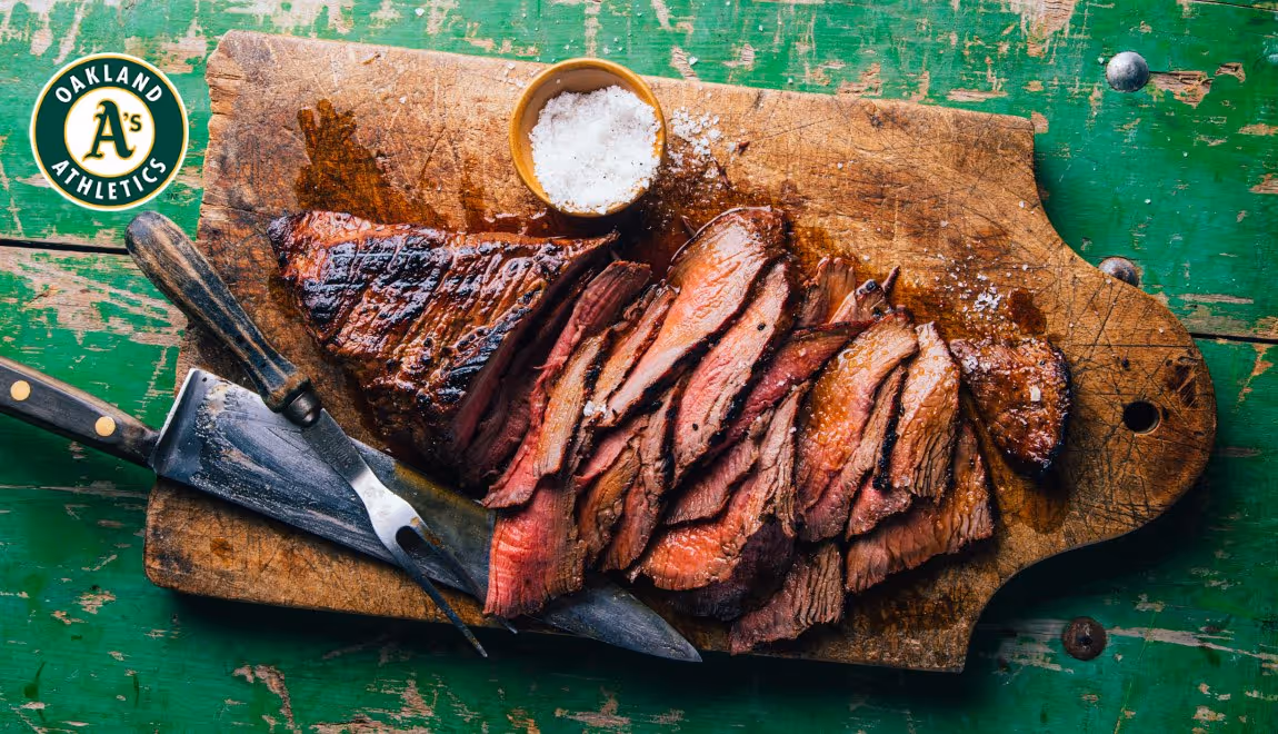 Sliced grilled steak on a wooden cutting board with a knife, fork, and a small bowl of coarse salt.