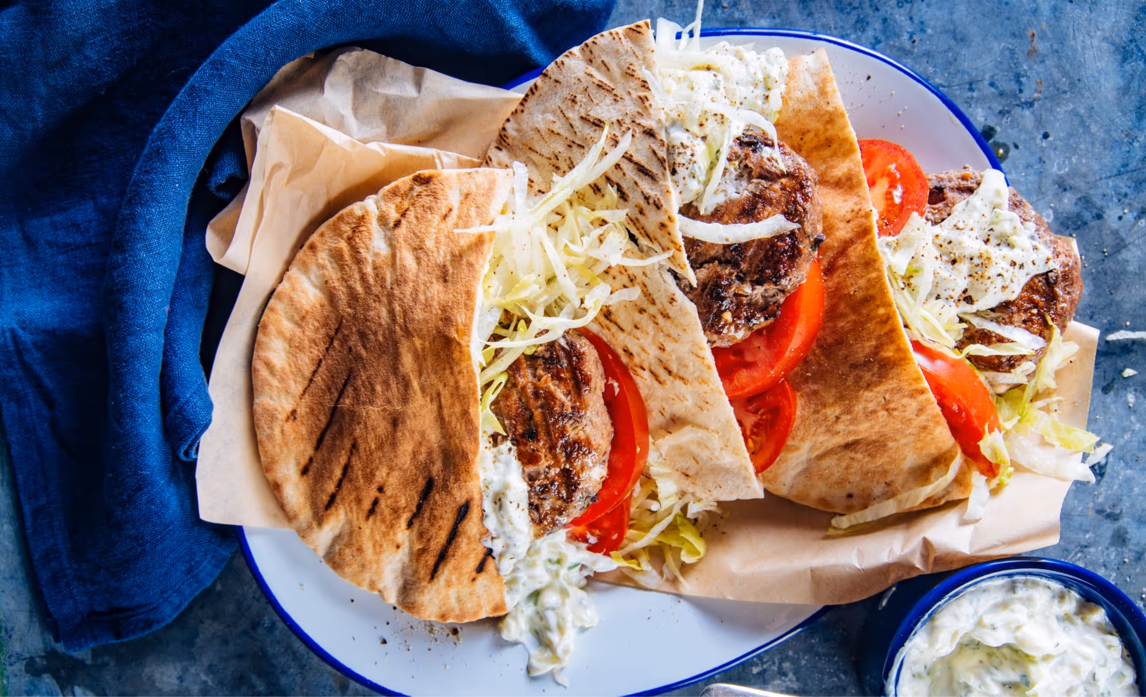 Three grilled pita sandwiches filled with meat patties, sliced tomatoes, shredded lettuce, and a creamy white sauce on a white plate with a blue rim, next to a blue cloth and a bowl of creamy sauce.