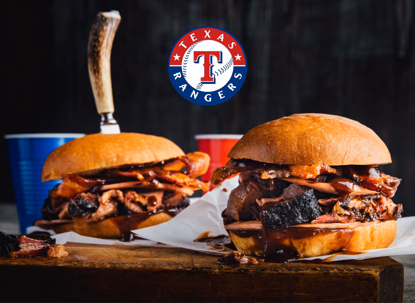 Two barbeque sandwiches with smoked meat and bacon on wooden board, with Texas Rangers logo above and red and blue plastic cups in the background.