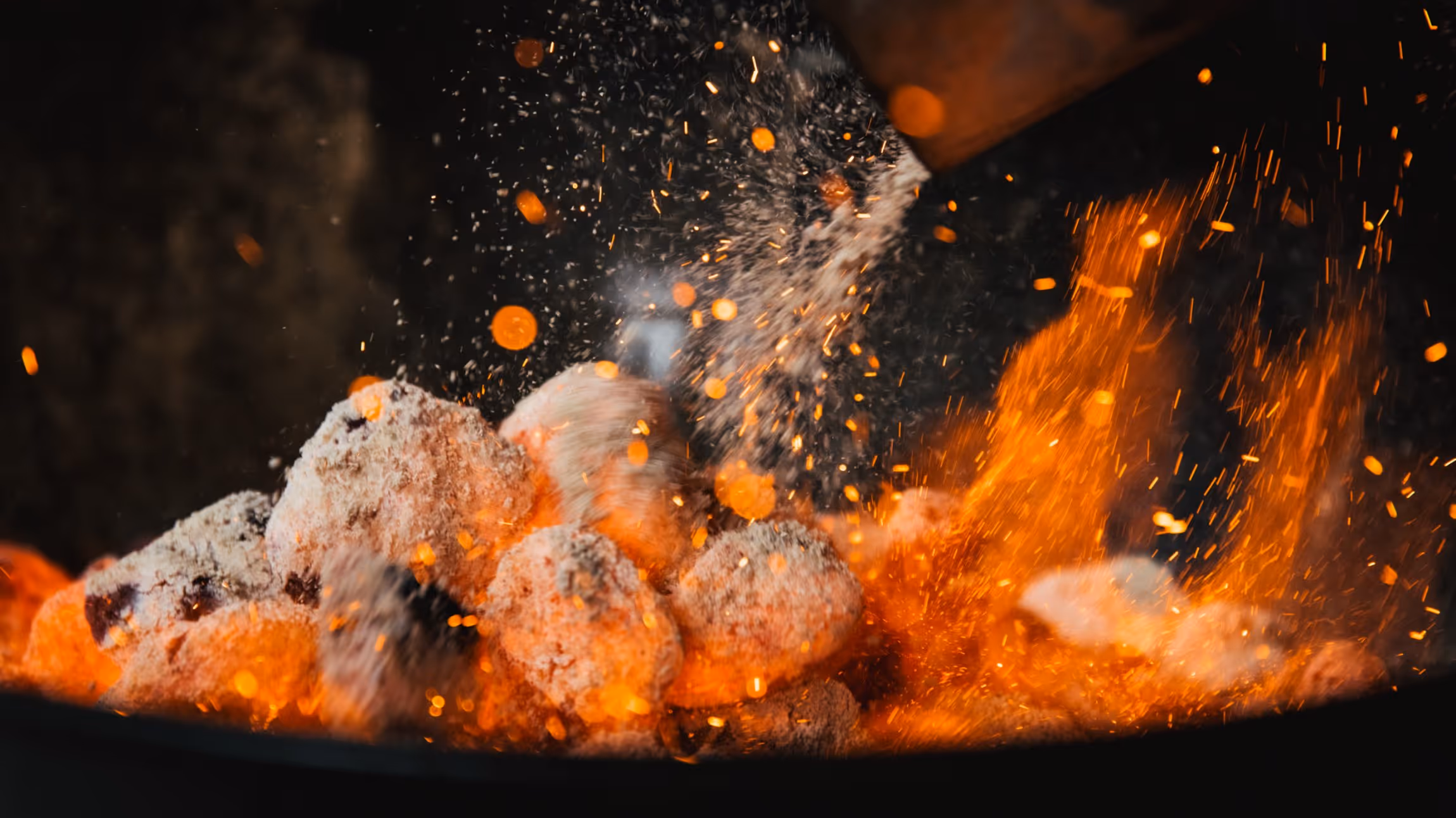 Glowing charcoal briquettes with sparks and ash flying in the air during grilling.