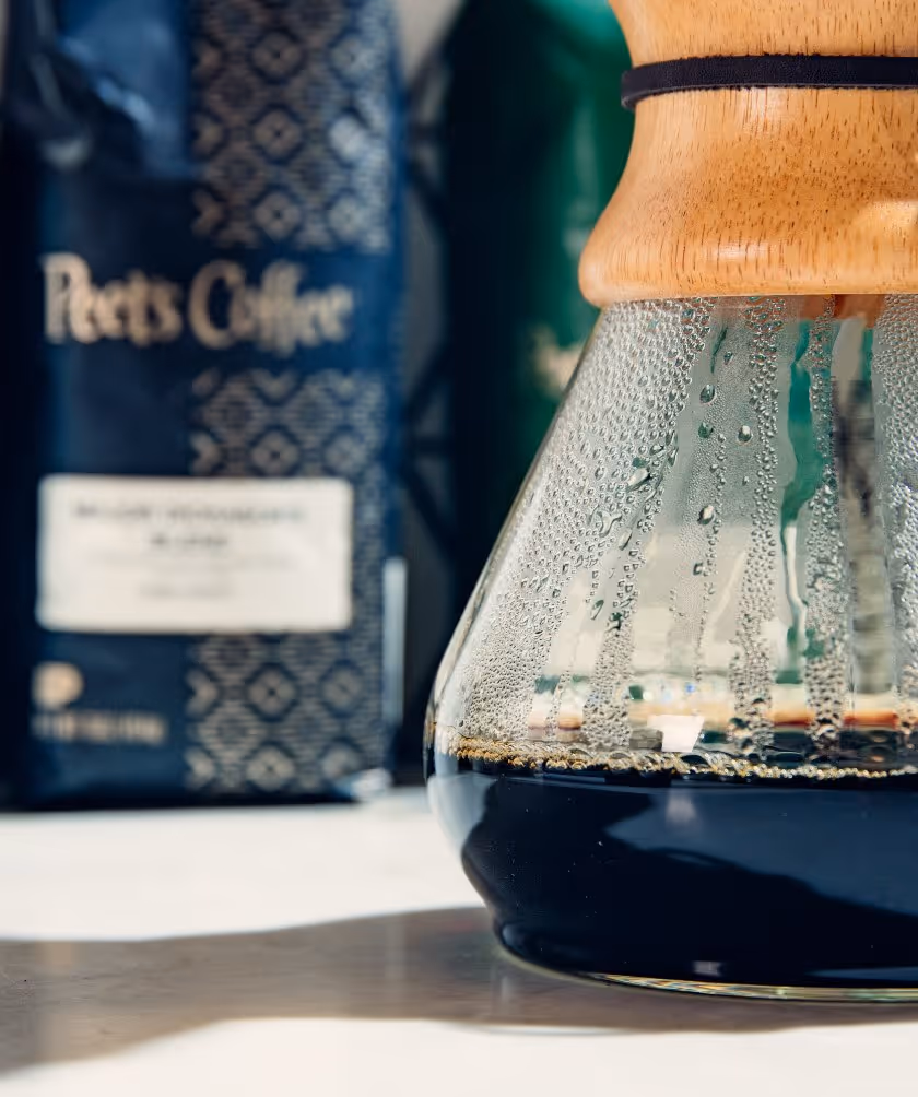 Close-up of a glass coffee carafe with condensation and dark brewed coffee, with a Peet's Coffee bag blurred in the background.