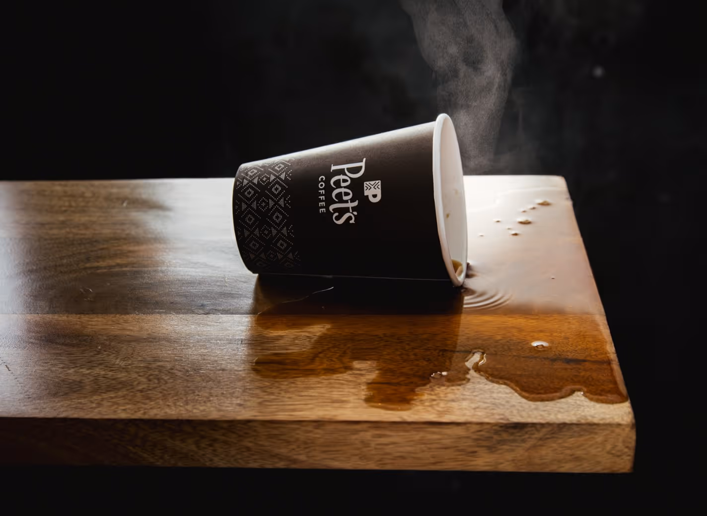 Spilled hot coffee from a tipped-over Peet's Coffee paper cup on a wooden table, with steam rising.