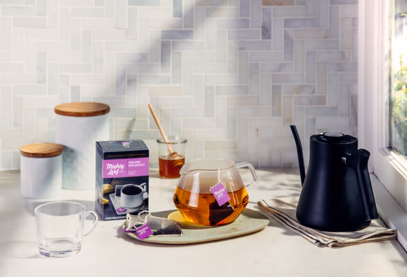 Glass teapot with amber tea and Mighty Leaf tea bag on a tray, next to a box of Mighty Leaf Organic Breakfast tea, empty glass cup, black kettle, and honey jar on a white kitchen counter.