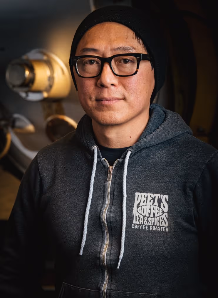 Man wearing black beanie, black glasses, and a dark gray hoodie with Peet's Coffee Tea & Spices logo standing indoors near coffee equipment.