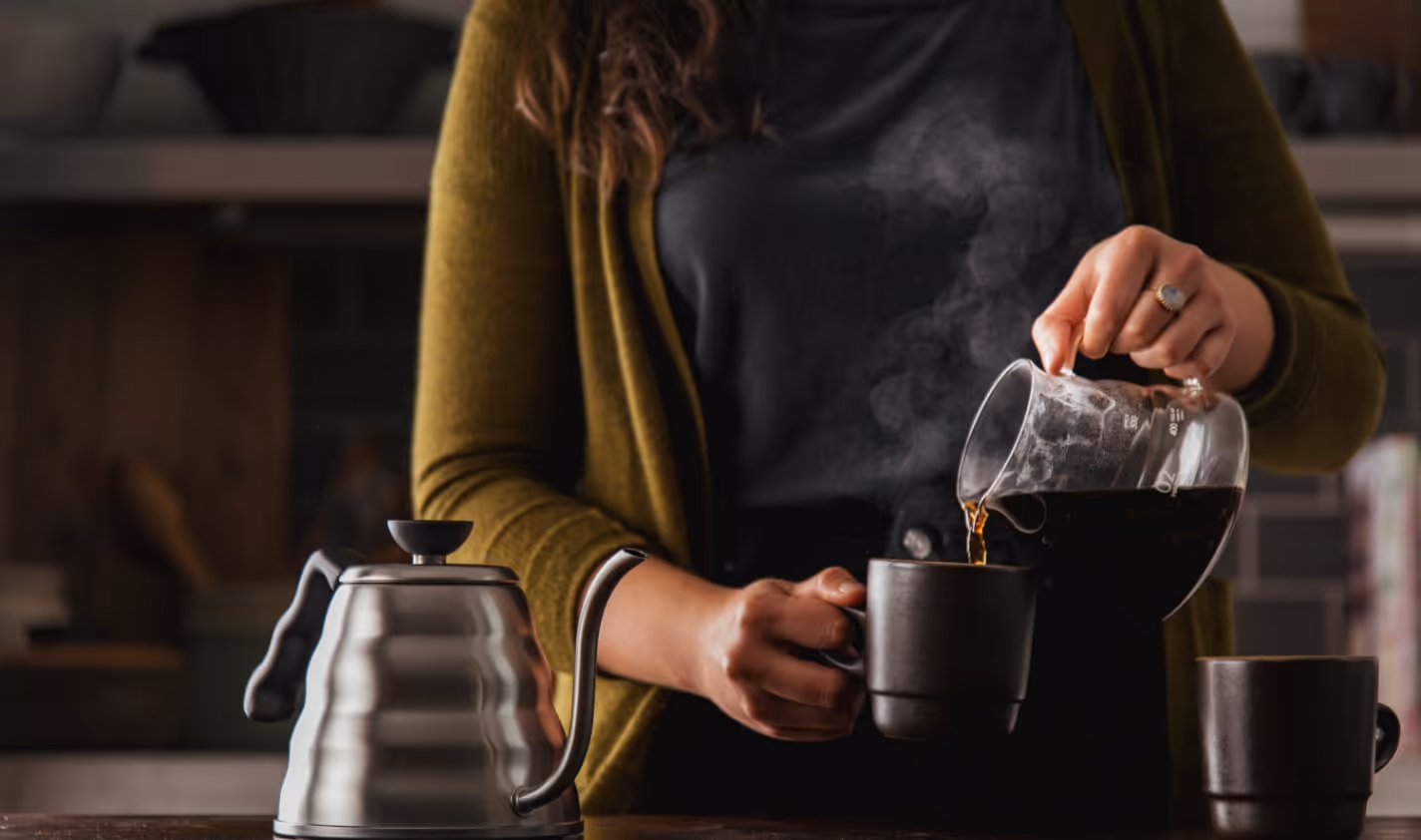 Person pouring hot coffee from a glass carafe into a black mug with a stainless steel kettle on the table.