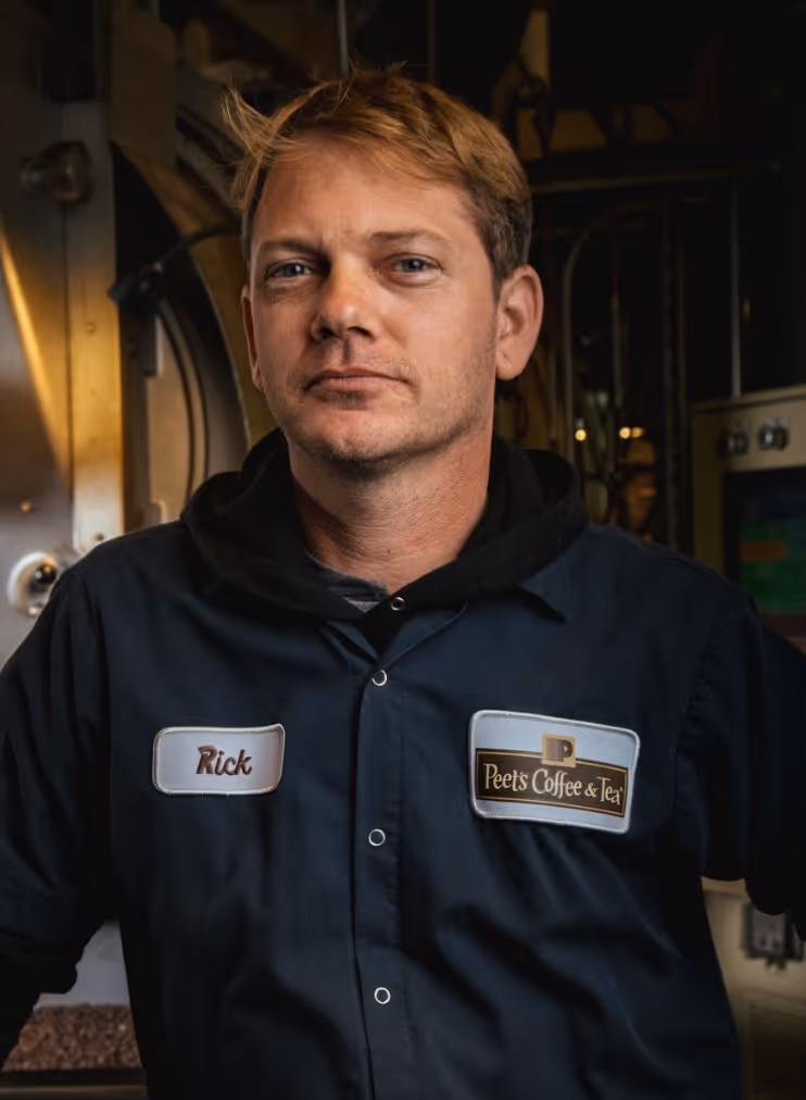 Man wearing a dark Peet's Coffee & Tea uniform with name tag 'Rick' in a dimly lit industrial setting.