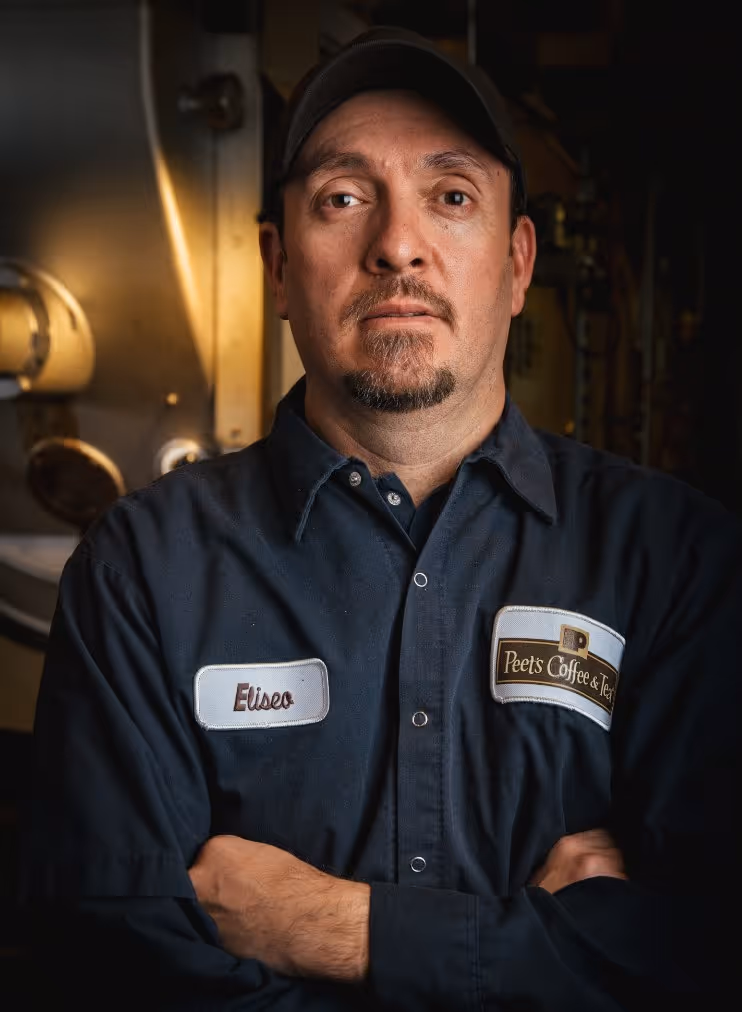 Man wearing a dark shirt and cap with name tag 'Eliseo' and a 'Peet's Coffee & Tea' logo patch, standing with arms crossed.