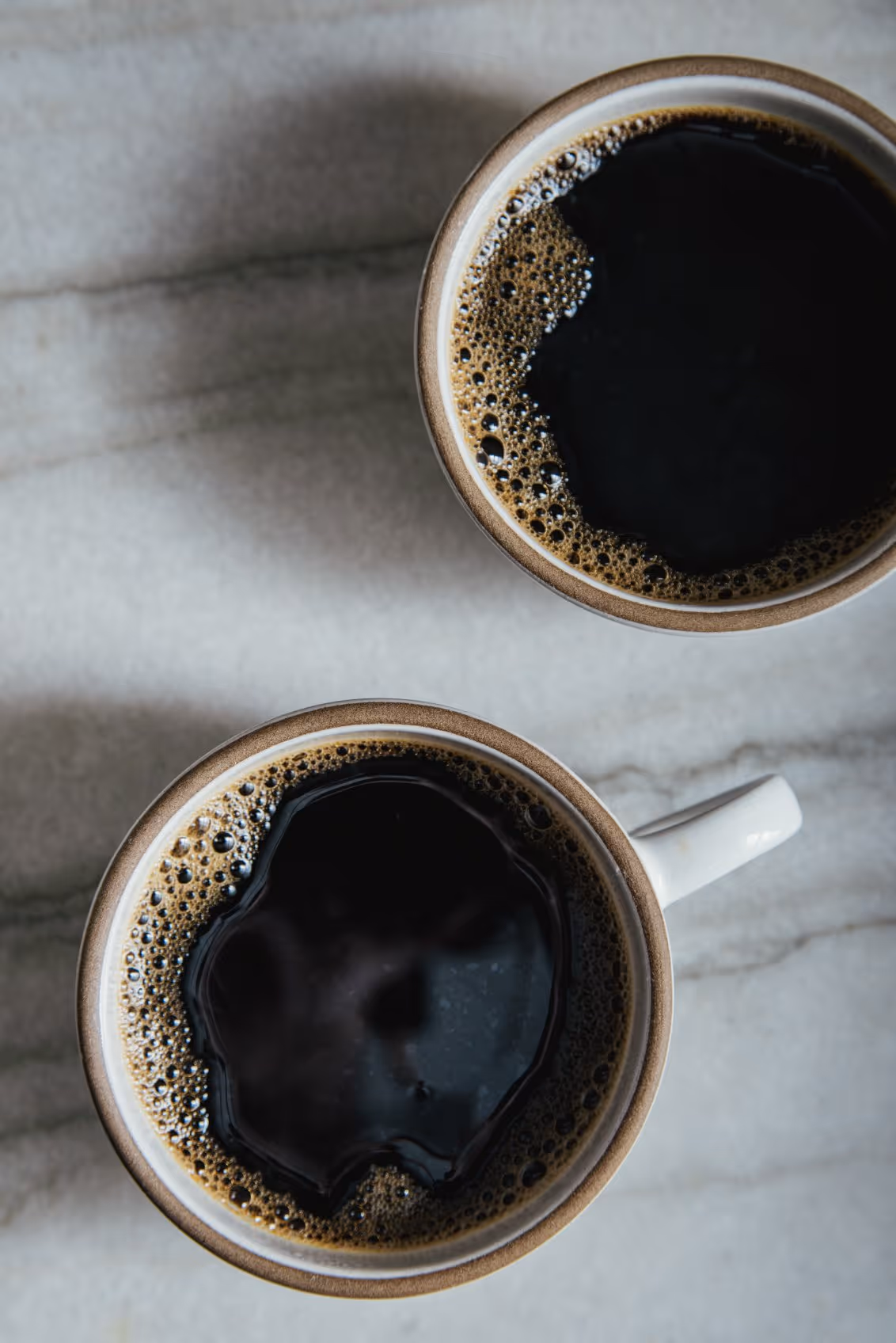 Top view of two cups of black coffee with bubbles on the surface on a marble countertop.