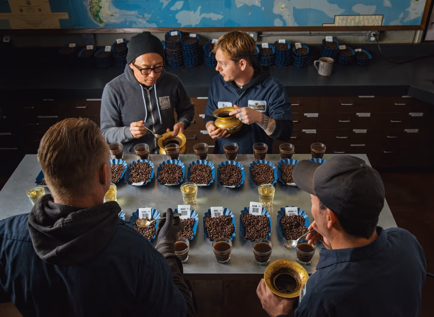 Four people at a table cupping and evaluating multiple blue trays of coffee beans and glasses of brewed coffee.