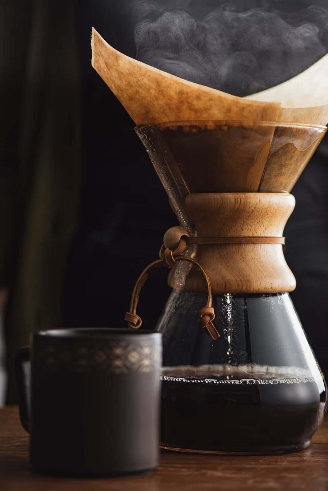 Close-up of a glass Chemex coffee maker with a wooden collar and steaming coffee, next to a dark ceramic mug on a wooden surface.