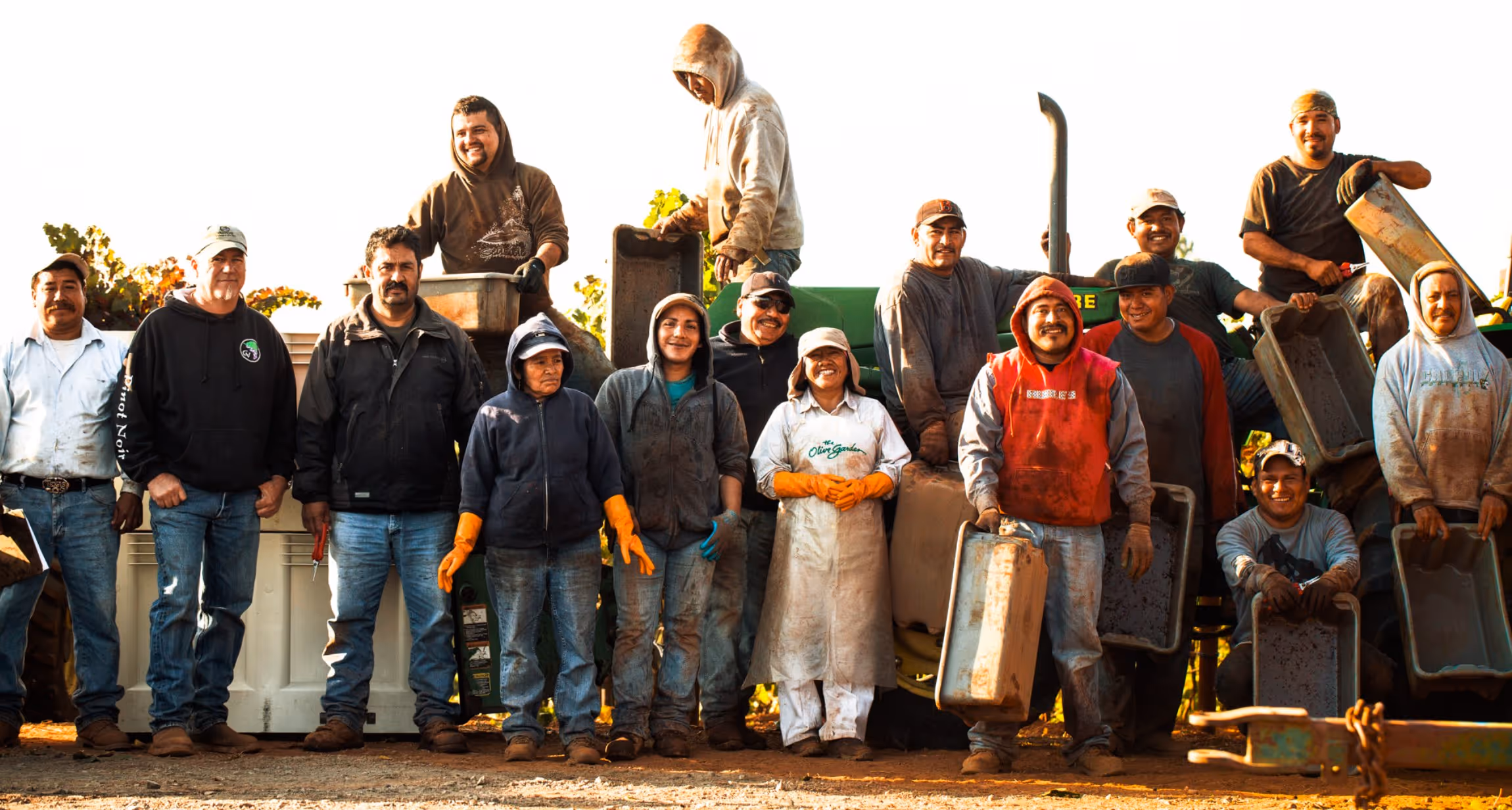 Group of farm workers wearing gloves and work clothes standing and sitting in front of agricultural equipment with grapevines in the background.