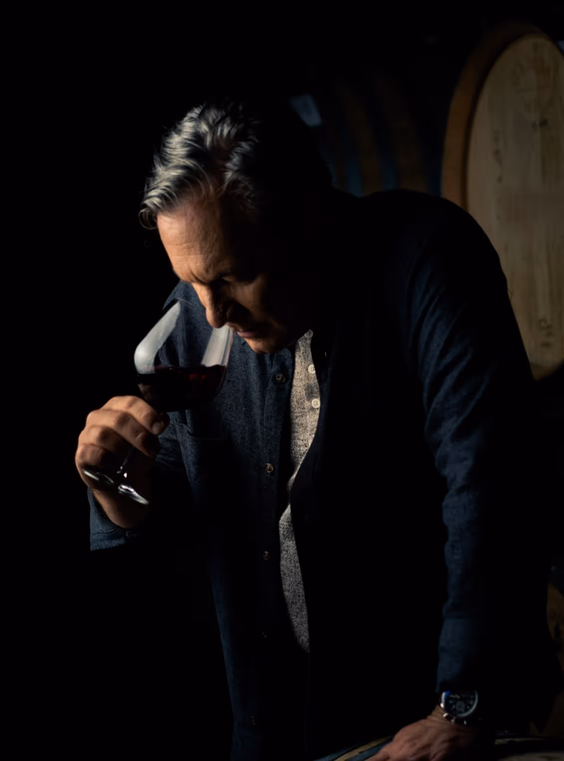 Man smelling red wine in a glass inside a dimly lit wine cellar with wooden barrels in the background.