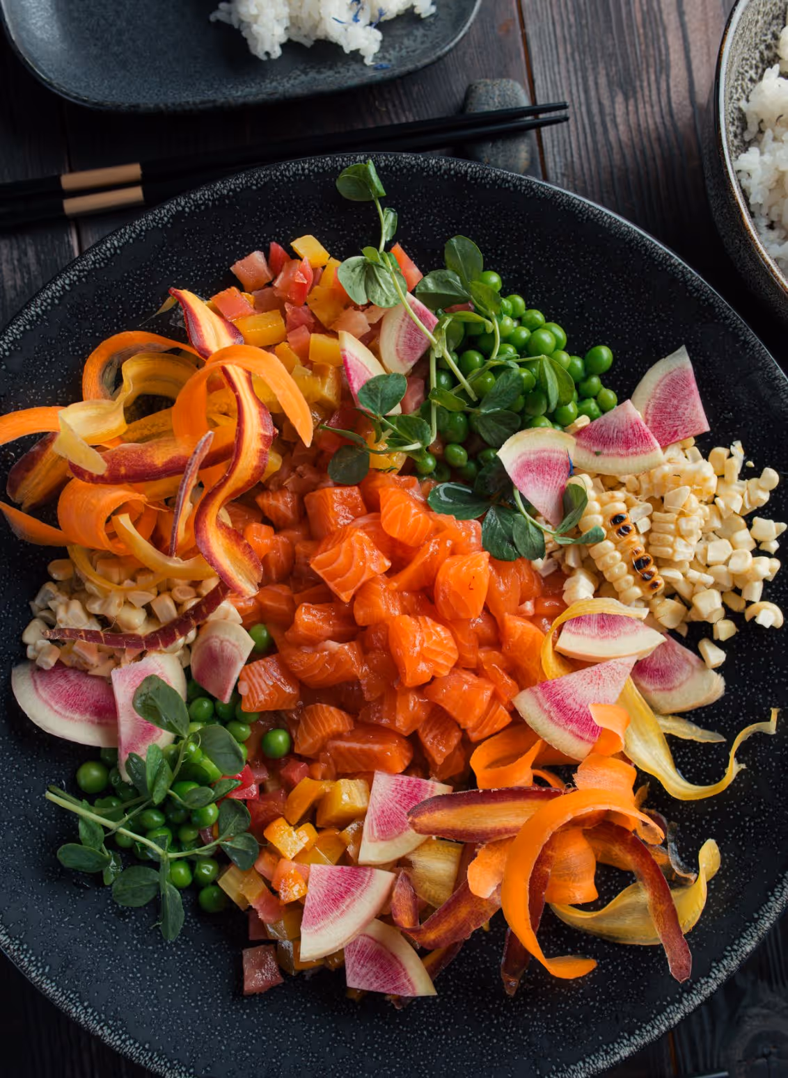Black plate with diced raw salmon surrounded by peas, corn, colorful vegetable curls, diced bell peppers, and watermelon radish slices.