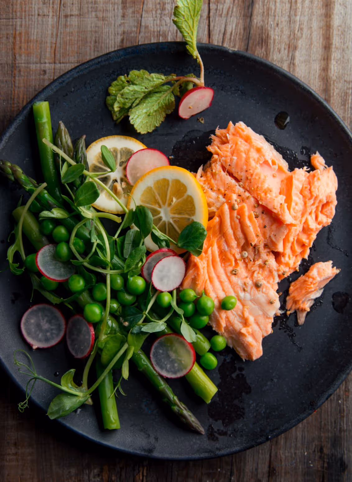 Black plate with cooked salmon, asparagus, green peas, radish slices, lemon slices, and fresh herbs on a wooden surface.