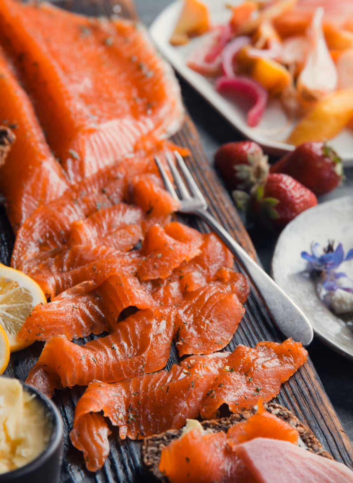Thin slices of smoked salmon on a wooden board with lemon wedges, fresh strawberries, and a fork.