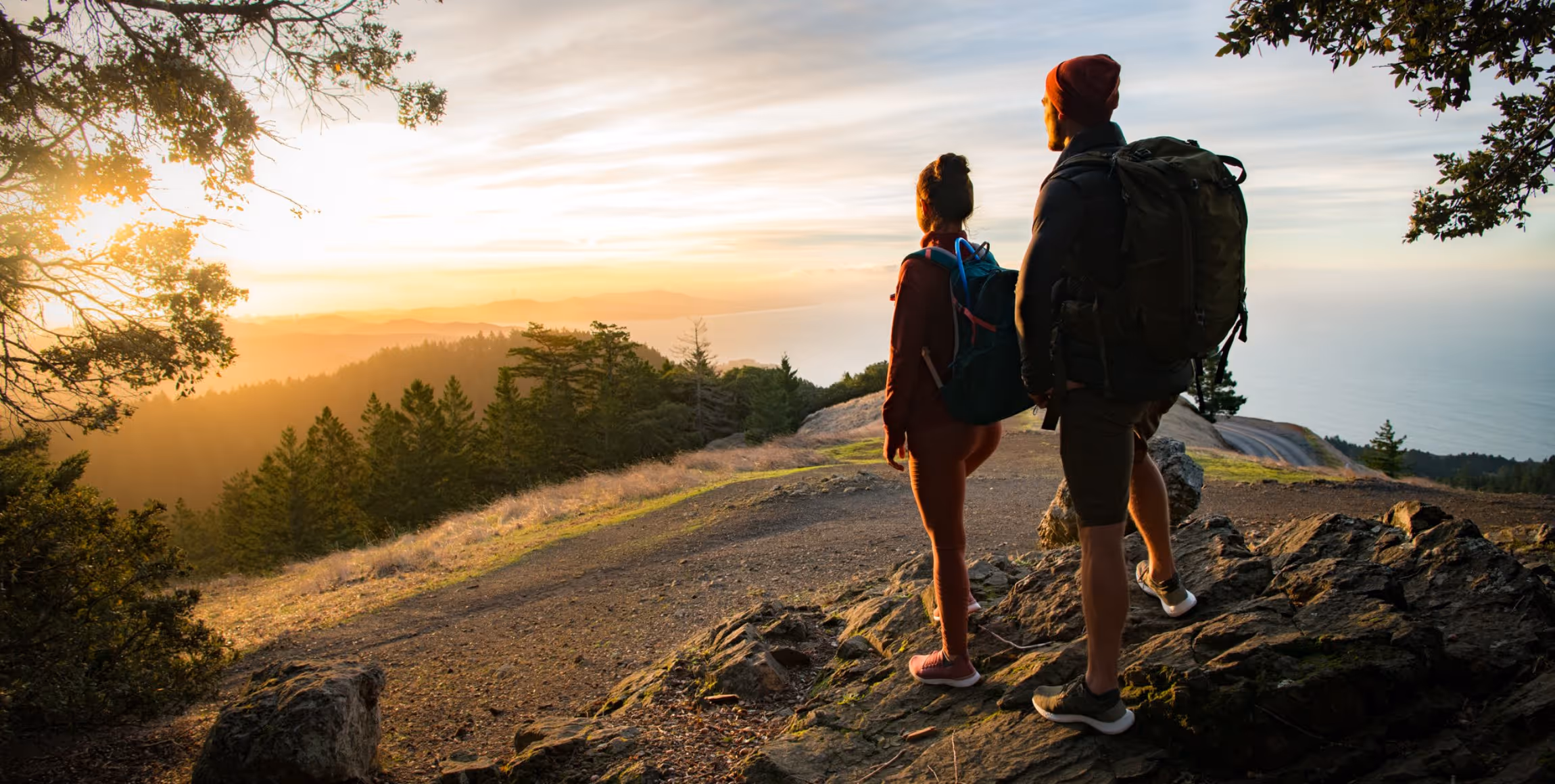 Two hikers standing on rocky terrain overlooking a forest and ocean at sunset.