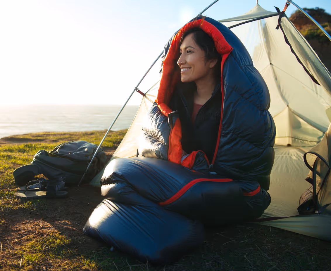 Smiling woman wrapped in a black and red sleeping bag sitting at the entrance of a tent near the coast at sunset.