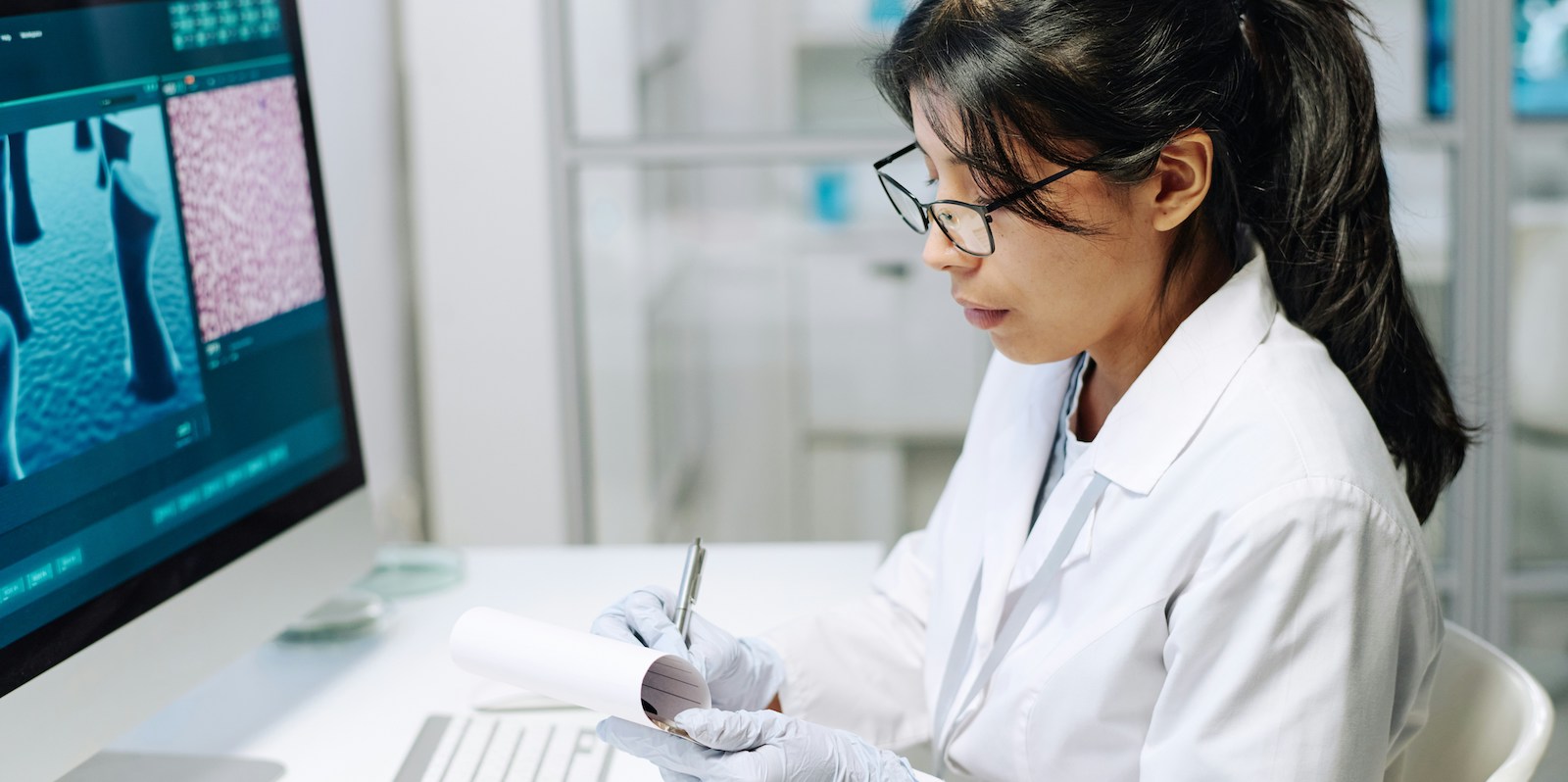 Scientist in a white lab coat and gloves writing notes with a pen at a workstation with a computer displaying microscopic images.