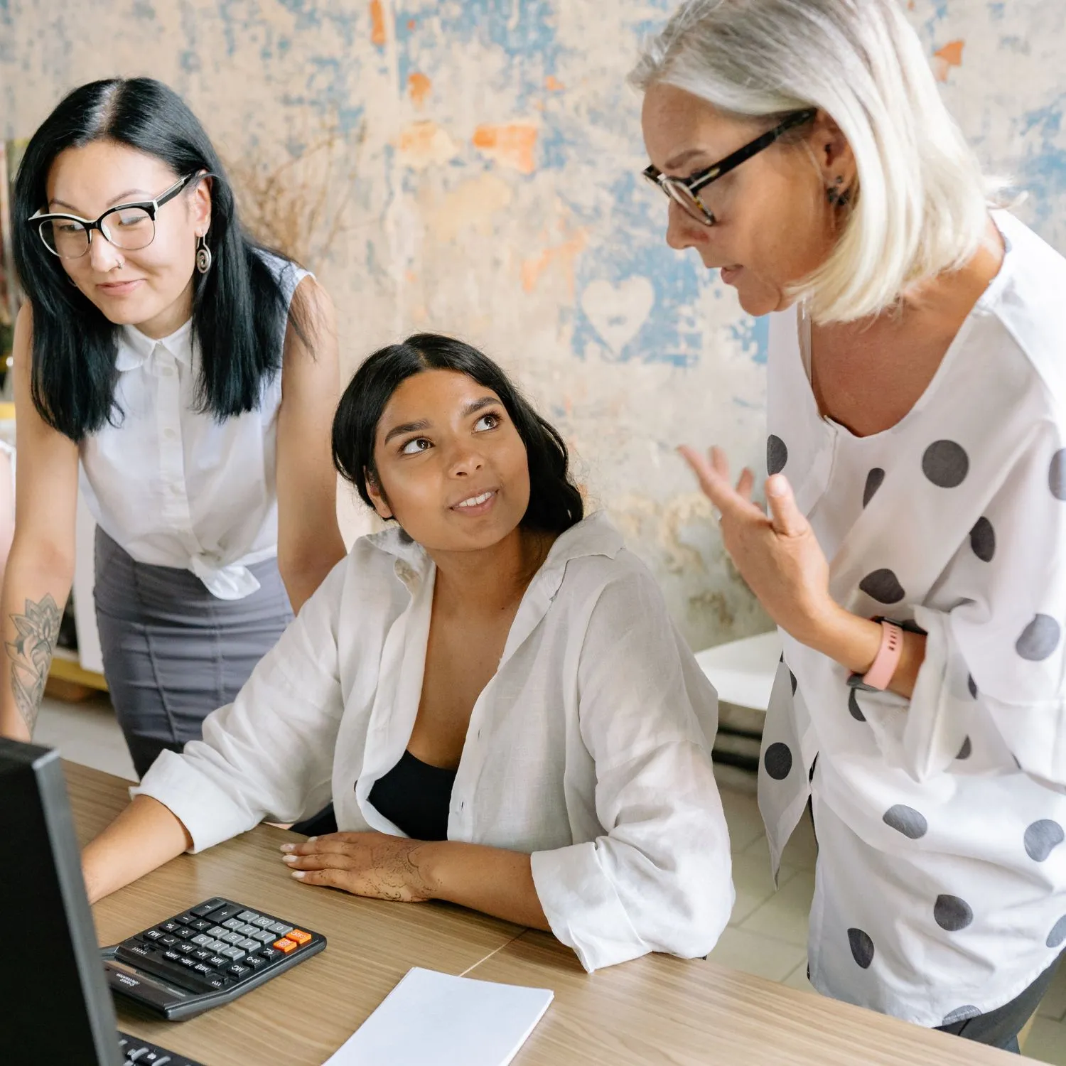Image representing people development via coaching for engagement and retention. Shows three women collaborating at a desk with a calculator, notebook, and desktop computer in an office.