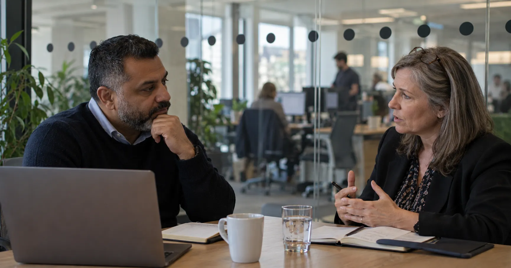 Two professionals engaged in a workplace leadership coaching session for development, with an open office scene in the background. 
